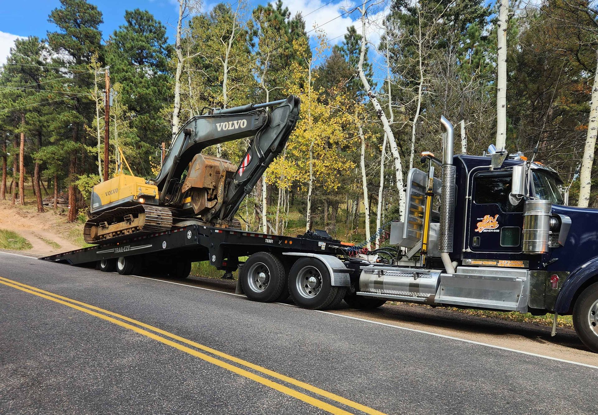 Semi truck hauling a yellow excavator on a lowboy trailer with aspens behind